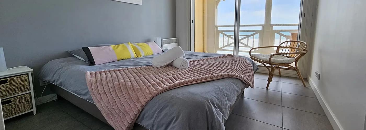 Appart hôtel Pasino n°6 chambre tons pastels, donnant sur la terrasse vue mer, 2 chevets, 2 lampes de chevet, 2 coussins décoration sur le lit, un plaid rose poudré et 2 serviettes de bain blanches, décorations au mur et plafond, un fauteuil en rotin et son coussin blanc. A l'arrière, la terrasse donnant sur l'océan
