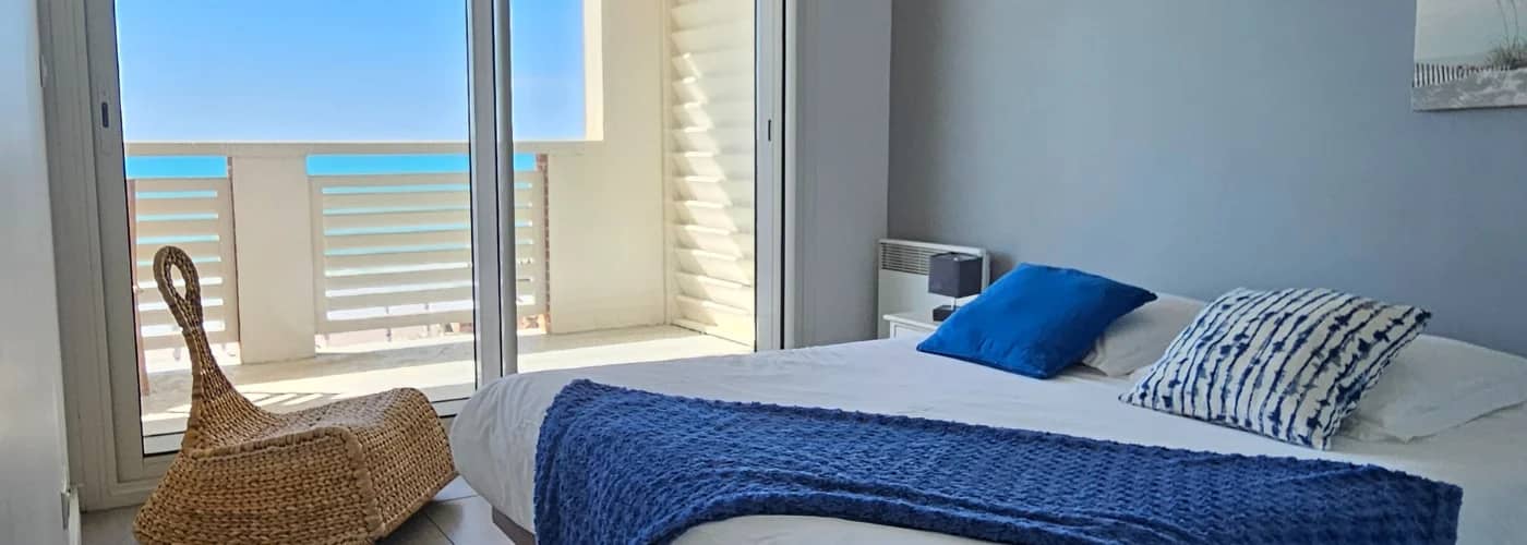 appart hôtel pasino n°5, chambre donnant sur la terrasse vue mer, 2 lampes de chevet, 2 coussins décoration, 1 plaid posé sur le lit. Décorations au mur et plafond. A l'arrière de la photo vue sur la terrasse et l'océan