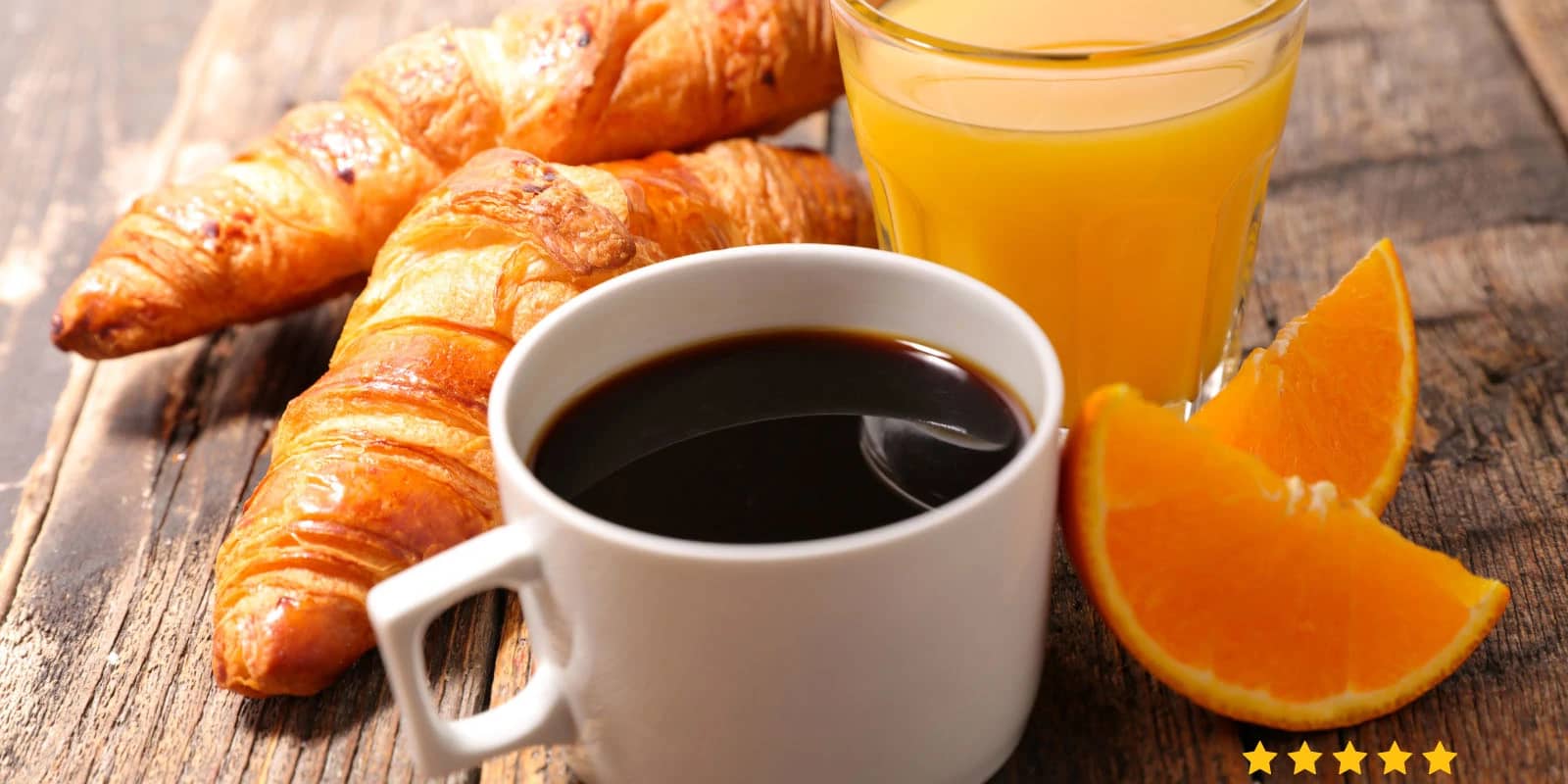 petit-dejeuner Petit dejeuner avec en premier plan une tasse blanche grand format contenant du café, et derrière on voit, posés sur une table en bois, 2 croissants. A droite des croissants, un grand verre de jus d'orange et deux quartiers d'orange.