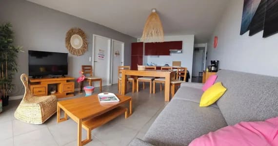 Appart hôtel Pasino N°1,pièce à vivre Au fond, la kitchenette équipée rouge, au milieu une table en teck et ses chaises, le long du mur à droite, une console en teck sur laquelle sont posés une cafetière, un grille pain et une bouilloire, à l'avant à droite un canapé et ses coussins. Au mur un tableau. Sur la gauche à l'avant un fauteuil en rotin, une table basse en teck et le long du mur, un palmier, un meuble TV et sa TV grand écran. Sur le mur et autres une décoration exotique